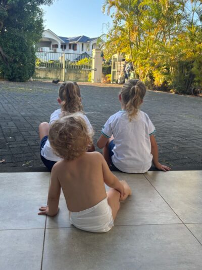 Expatriation en famille à Maurice : un quotidien rythmé par l’école et la douceur de vivre Trois jeunes enfants assis devant leur maison à l’île Maurice, attendant le bus scolaire dans un cadre tropical verdoyant.